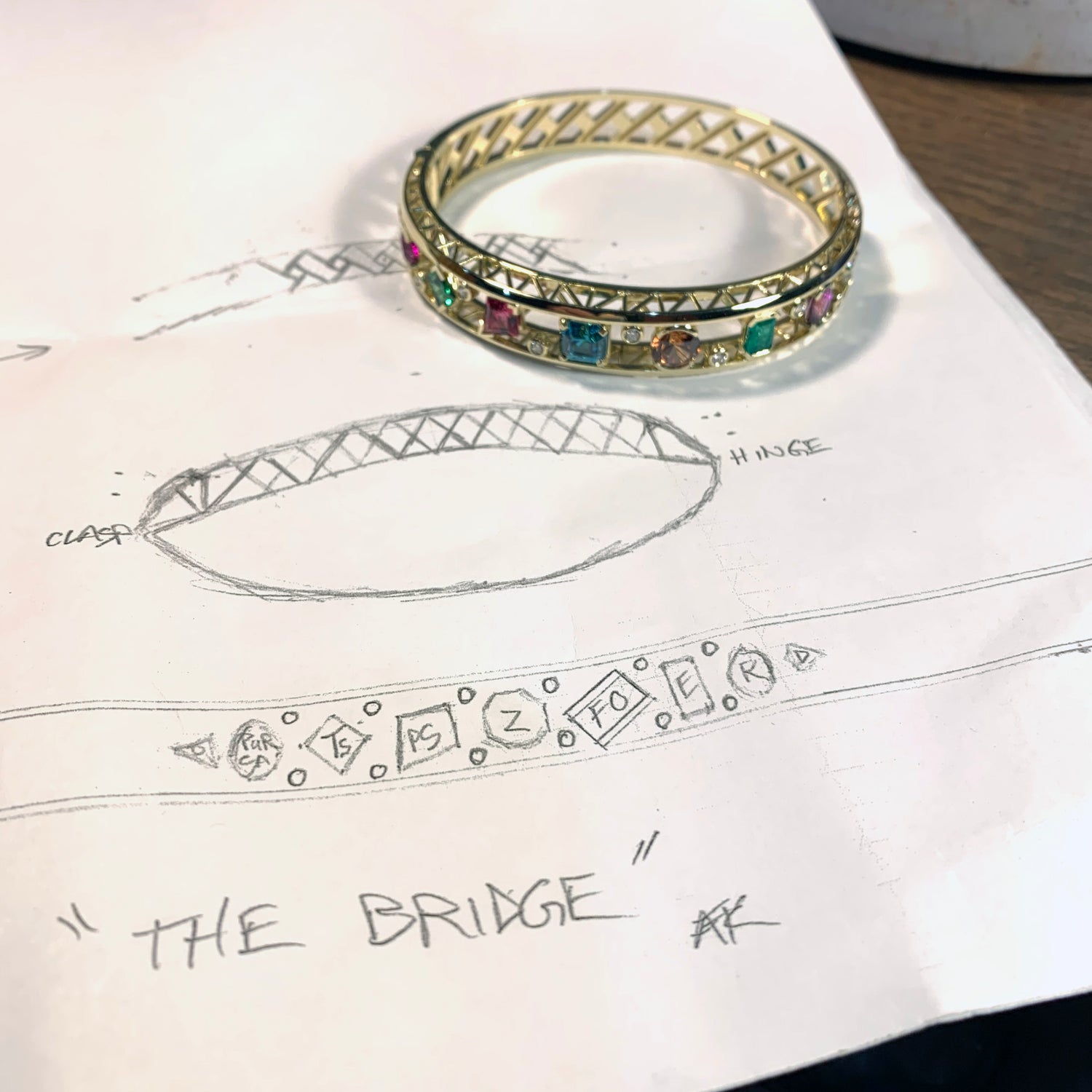 several sketches on a piece of paper with a bracelet design are shown with the finished design sitting on top of the paper. The 18k yellow gold bracelet is filled with brightly colored gemstones and diamonds and is titled "the bridge"