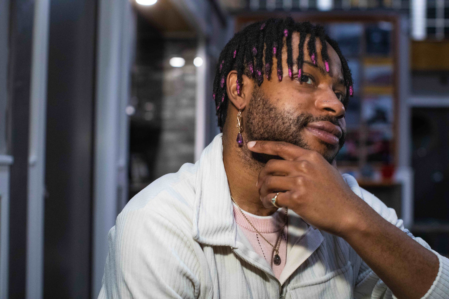 An African American man wearing jewelry created at Glitter and Gold looks into the distance and holding his hand to his chin.  He is wearing dangly earrings, a ring with a sapphire, and a pendant.