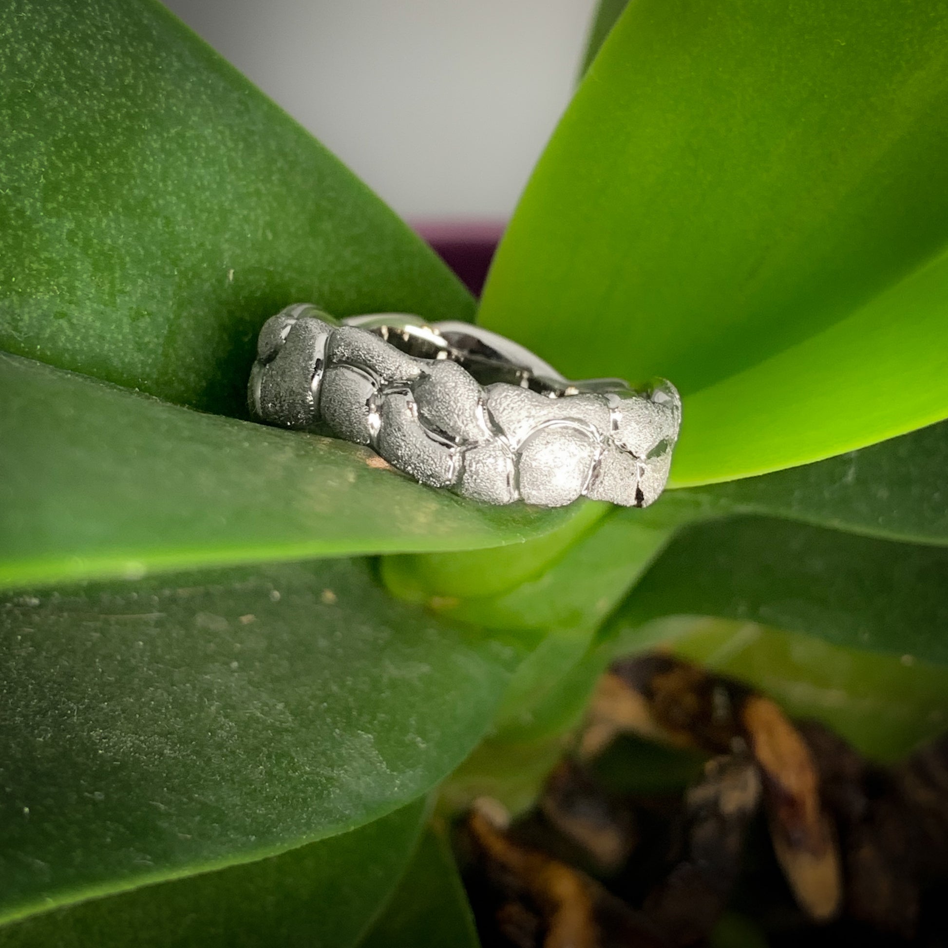 A white gold ring with moss like texture on a double layer of rocks carved into the ring.  The ring has a meandering edge and has shiny paths between the "rocks".   The ring is nestled into an orchid plant.